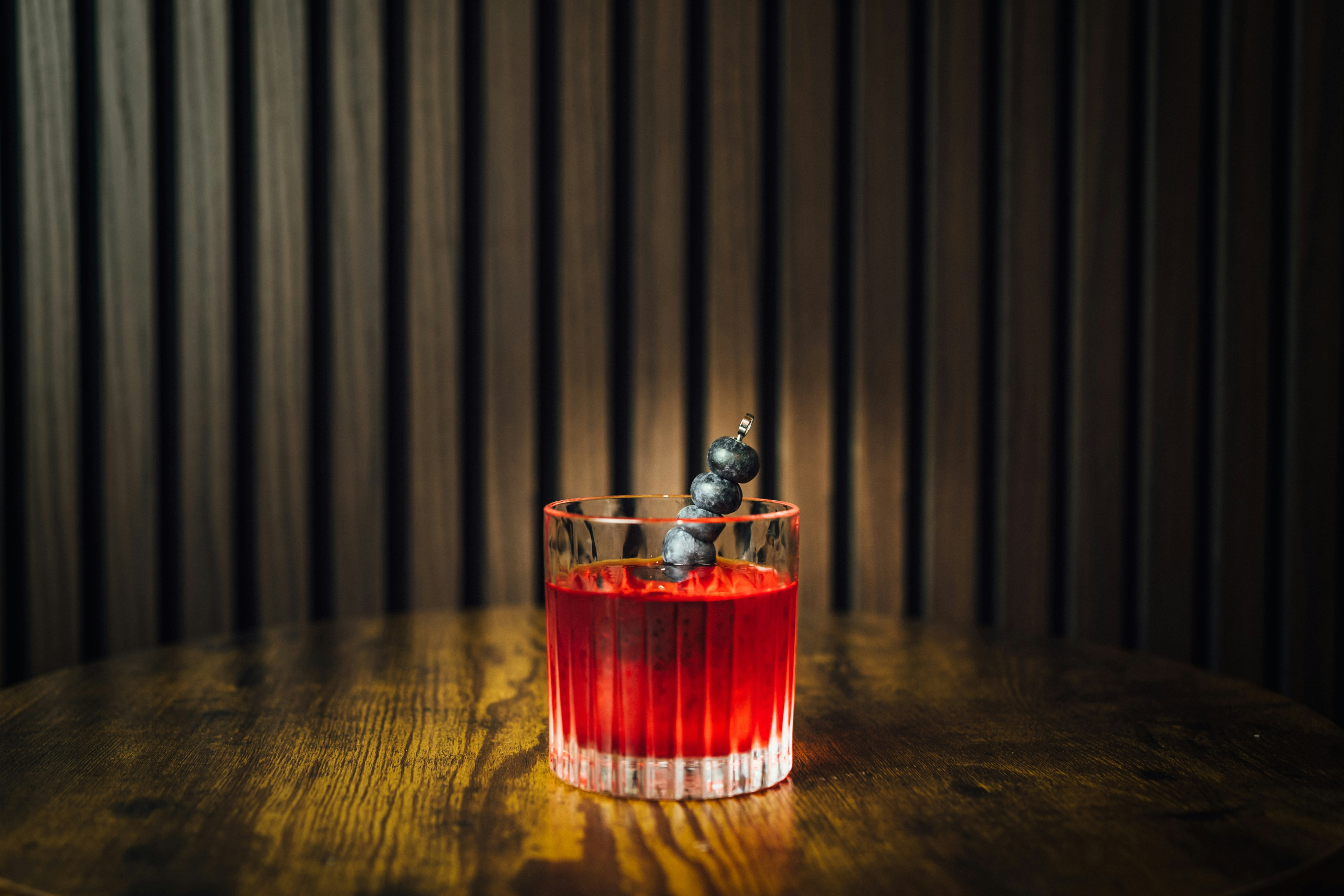 Image of a small glass on a wooden table filled with a redish liquid and garnished with a cocktail stick through three blueberries.